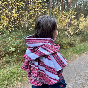 May include: A white and red embroidered hooded scarf with a geometric pattern. The scarf is layered and has a decorative trim. The person is wearing a dark blue embroidered top. The background is a forest with trees and yellow leaves.