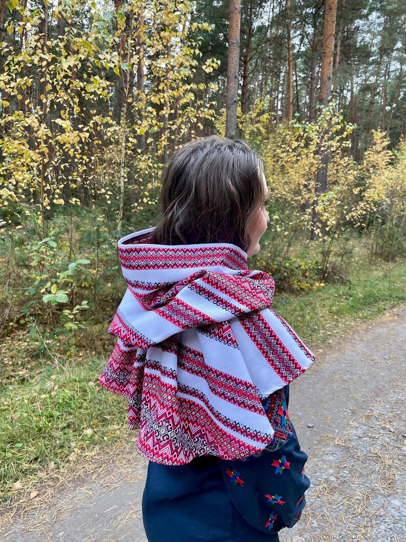 May include: A white and red embroidered hooded scarf with a geometric pattern. The scarf is layered and has a decorative trim. The person is wearing a dark blue embroidered top. The background is a forest with trees and yellow leaves.