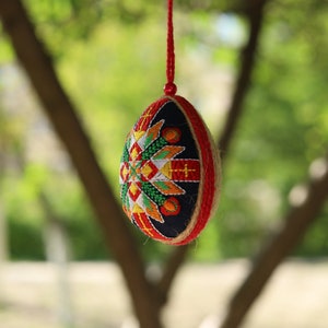 K&ouml;nnte beinhalten: Ein rotes, wei&szlig;es und blaues besticktes Ei-Ornament, das an einer roten Schnur h&auml;ngt. Das Ei ist mit einem geometrischen Muster verziert und von einem gr&uuml;nen, verschwommenen Hintergrund umgeben.