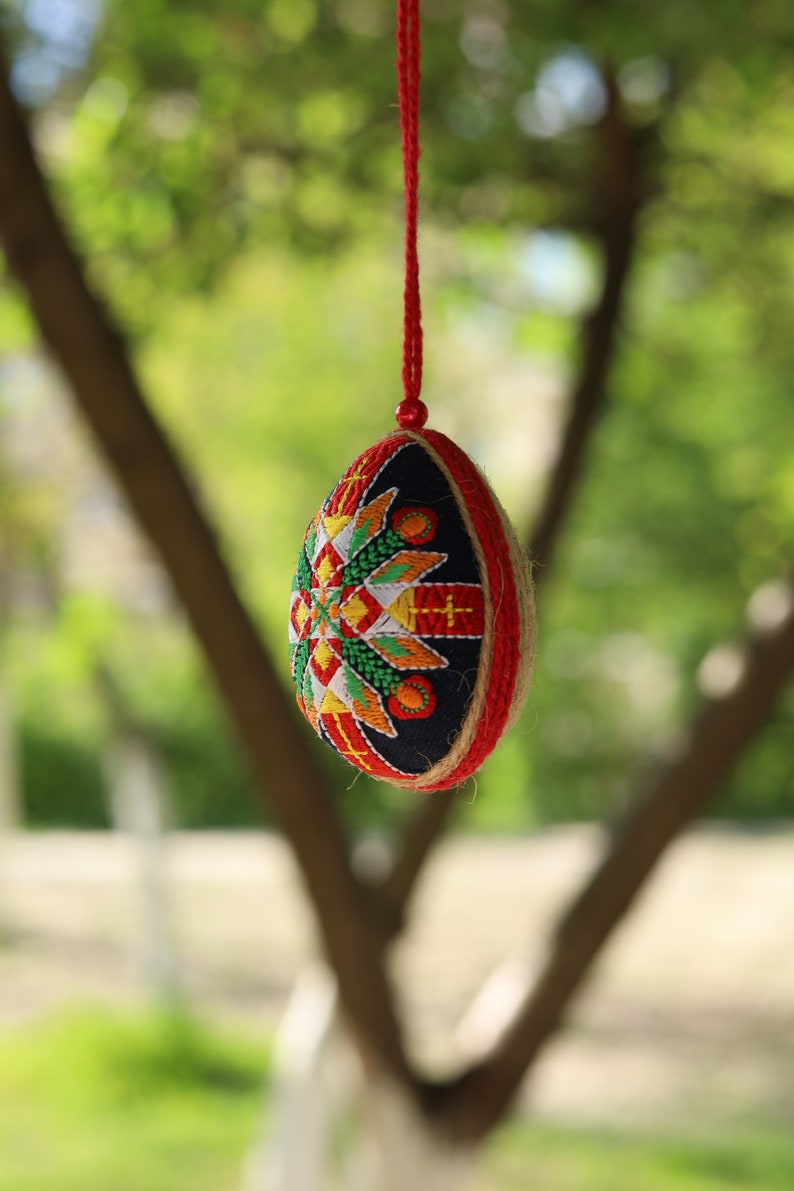 K&ouml;nnte beinhalten: Ein rotes, wei&szlig;es und blaues besticktes Ei-Ornament, das an einer roten Schnur h&auml;ngt. Das Ei ist mit einem geometrischen Muster verziert und von einem gr&uuml;nen, verschwommenen Hintergrund umgeben.