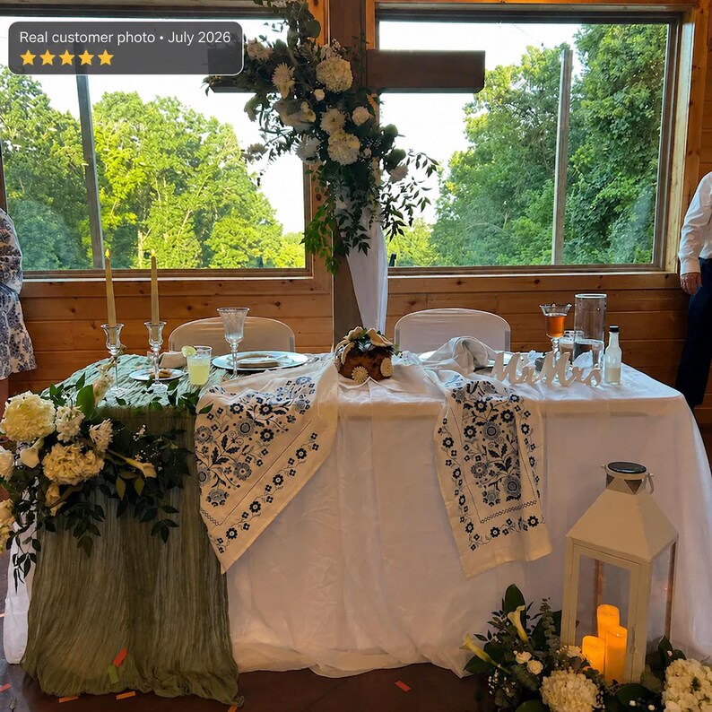 May include: A wedding sweetheart table covered with a white tablecloth and a sage green runner, featuring a white table runner with blue floral embroidery, white floral arrangements, two tall candles, glassware, and a white lantern on the floor.