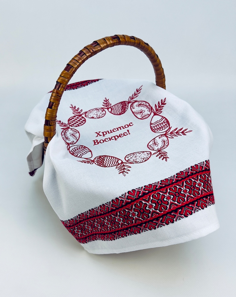 May include: White fabric Easter basket with a brown wicker handle. The fabric has a red embroidered border and a wreath of red embroidered eggs with the text "Христос Воскрес!" in the center.
