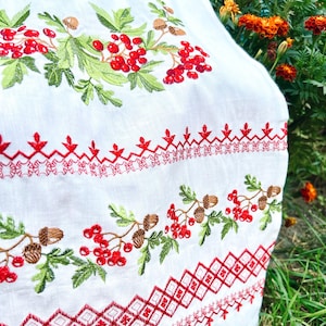 K&ouml;nnte beinhalten: Wei&szlig;er Stoff mit detaillierter Stickerei mit roten Beeren, gr&uuml;nen Bl&auml;ttern und braunen Eicheln. Das Design umfasst dekorative rote N&auml;hte in geometrischen und floralen Mustern und schafft eine Volkskunst-&Auml;sthetik.