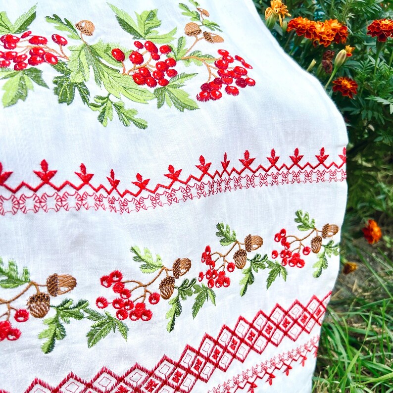 K&ouml;nnte beinhalten: Wei&szlig;er Stoff mit detaillierter Stickerei mit roten Beeren, gr&uuml;nen Bl&auml;ttern und braunen Eicheln. Das Design umfasst dekorative rote N&auml;hte in geometrischen und floralen Mustern und schafft eine Volkskunst-&Auml;sthetik.