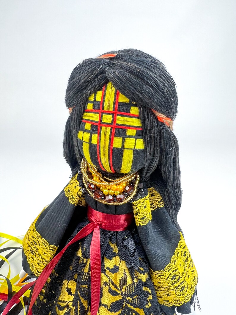 K&ouml;nnte beinhalten: Handgemachte Puppe mit schwarzen Garnhaaren und einem gelb-rot gemusterten Gesicht. Die Puppe tr&auml;gt ein schwarzes Kleid mit gelbem Spitzensaum und einem roten Band. Eine Perlenkette ziert den Hals der Puppe.