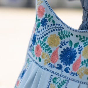 May include: Close-up of a light blue garment with intricate floral embroidery. The design features blue, yellow, and coral flowers with green leaves. The edges are finished with a detailed blue stitch.