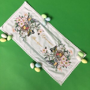 以下が含まれることがあります： 鳥、巣、花の刺繍が施された白いイースターテーブルランナー。 ランナーは装飾的なボーダーと「Christos Voskres」のテキストが特徴です。 カラフルなイースターエッグがランナーを囲んでいます。
