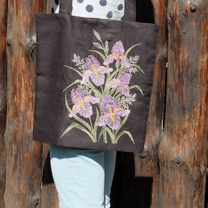 May include: A brown canvas tote bag featuring an embroidered design of purple iris flowers with yellow centres and green leaves.