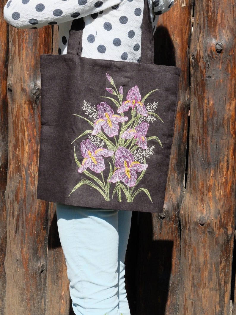 May include: A brown canvas tote bag featuring an embroidered design of purple iris flowers with yellow centres and green leaves.