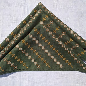 May include: Three folded green cloth napkins with embroidered diagonal patterns in gold and off-white. The napkins are arranged on a white tablecloth with a subtle geometric design, creating a contrast of textures and colors.
