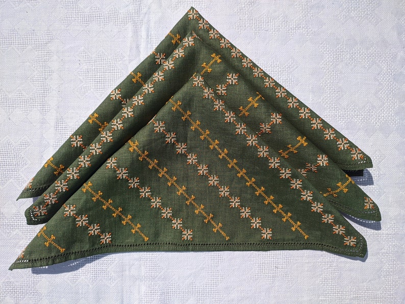 May include: Three folded green cloth napkins with embroidered diagonal patterns in gold and off-white. The napkins are arranged on a white tablecloth with a subtle geometric design, creating a contrast of textures and colors.