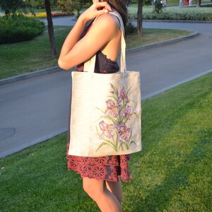 May include: A white canvas tote bag with a floral design featuring purple irises. The bag has long straps and is being held by a person.