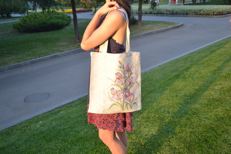 May include: A white canvas tote bag with a floral design featuring purple irises. The bag has long straps and is being held by a person.