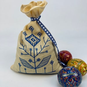 Puede incluir: Una bolsa de lino beige con cord&oacute;n y un dise&ntilde;o floral bordado en azul, atada con una cinta azul y blanca. Tres huevos de Pascua decorativos pintados en rojo, amarillo y azul est&aacute;n junto a la bolsa.