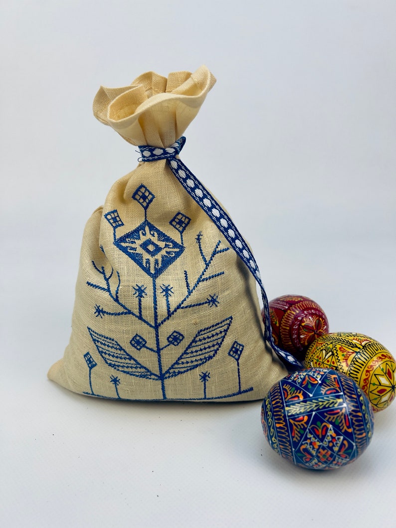 Puede incluir: Una bolsa de lino beige con cord&oacute;n y un dise&ntilde;o floral bordado en azul, atada con una cinta azul y blanca. Tres huevos de Pascua decorativos pintados en rojo, amarillo y azul est&aacute;n junto a la bolsa.