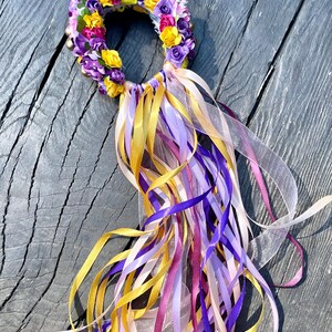 Puede incluir: Una corona de flores morada, amarilla y rosa con cuentas doradas y largos lazos de cinta morada, amarilla, rosa y blanca.