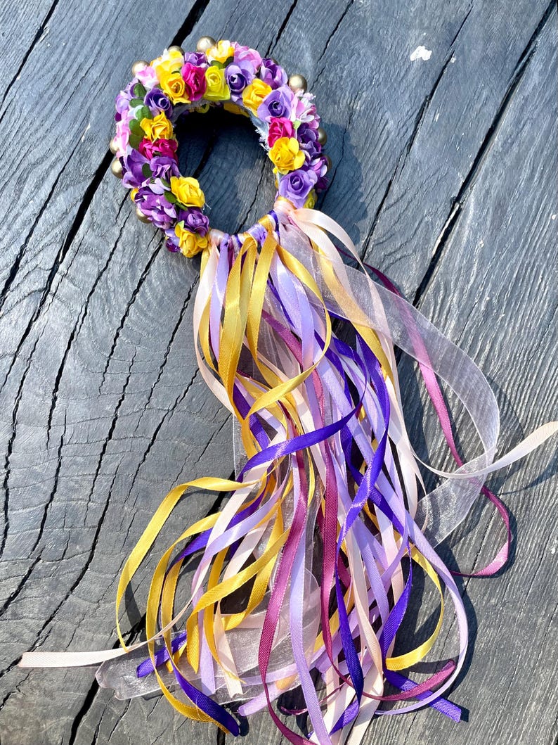 Puede incluir: Una corona de flores morada, amarilla y rosa con cuentas doradas y largos lazos de cinta morada, amarilla, rosa y blanca.
