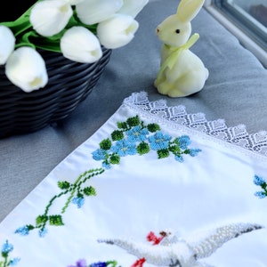 May include: White embroidered fabric featuring a floral design and a dove with an olive branch. The fabric is trimmed with white lace. A yellow ceramic bunny is in the background.