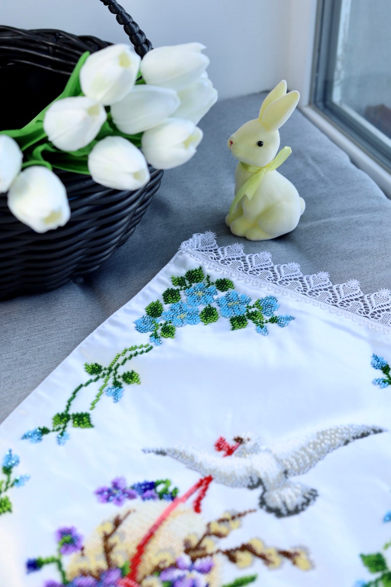 May include: White embroidered fabric featuring a floral design and a dove with an olive branch. The fabric is trimmed with white lace. A yellow ceramic bunny is in the background.