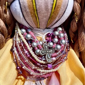 Puede incluir: Una mu&ntilde;eca hecha a mano con un vestido amarillo y marr&oacute;n, un cuello de encaje blanco y un colgante de cruz de plata. La mu&ntilde;eca lleva un collar hecho de m&uacute;ltiples hilos de cuentas en tonos rosa, morado y blanco.