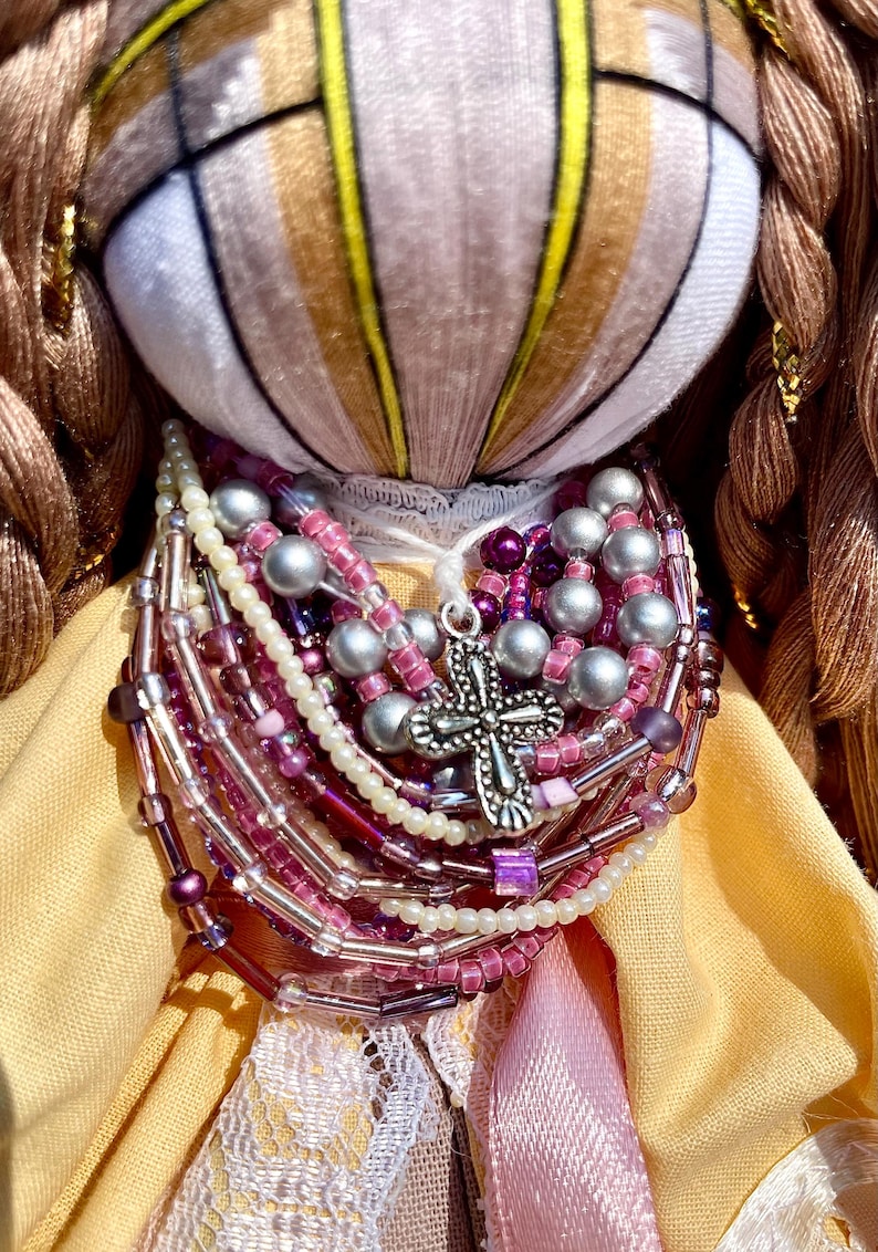 Puede incluir: Una mu&ntilde;eca hecha a mano con un vestido amarillo y marr&oacute;n, un cuello de encaje blanco y un colgante de cruz de plata. La mu&ntilde;eca lleva un collar hecho de m&uacute;ltiples hilos de cuentas en tonos rosa, morado y blanco.