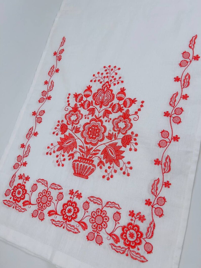 May include: White kitchen towel with red embroidered floral design. The design features a bouquet of flowers in a vase with a vine border.