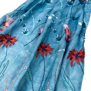 Op de afbeelding: Een lichtblauwe sjaal met geborduurde bloemenmotieven in tinten rood, roze en blauw.