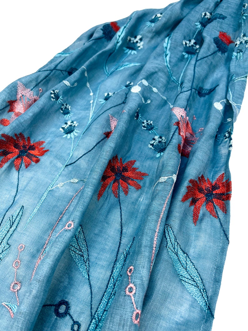 Op de afbeelding: Een lichtblauwe sjaal met geborduurde bloemenmotieven in tinten rood, roze en blauw.