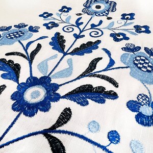 May include: Close-up of white fabric with intricate blue floral embroidery. The design features stylized flowers, leaves, and vines in shades of blue and black. The embroidery is detailed and creates a textured surface.