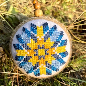 May include: A handmade Christmas ornament with a blue, yellow, and white geometric pattern. The ornament is made of burlap and is decorated with a blue and yellow cross-stitch design.