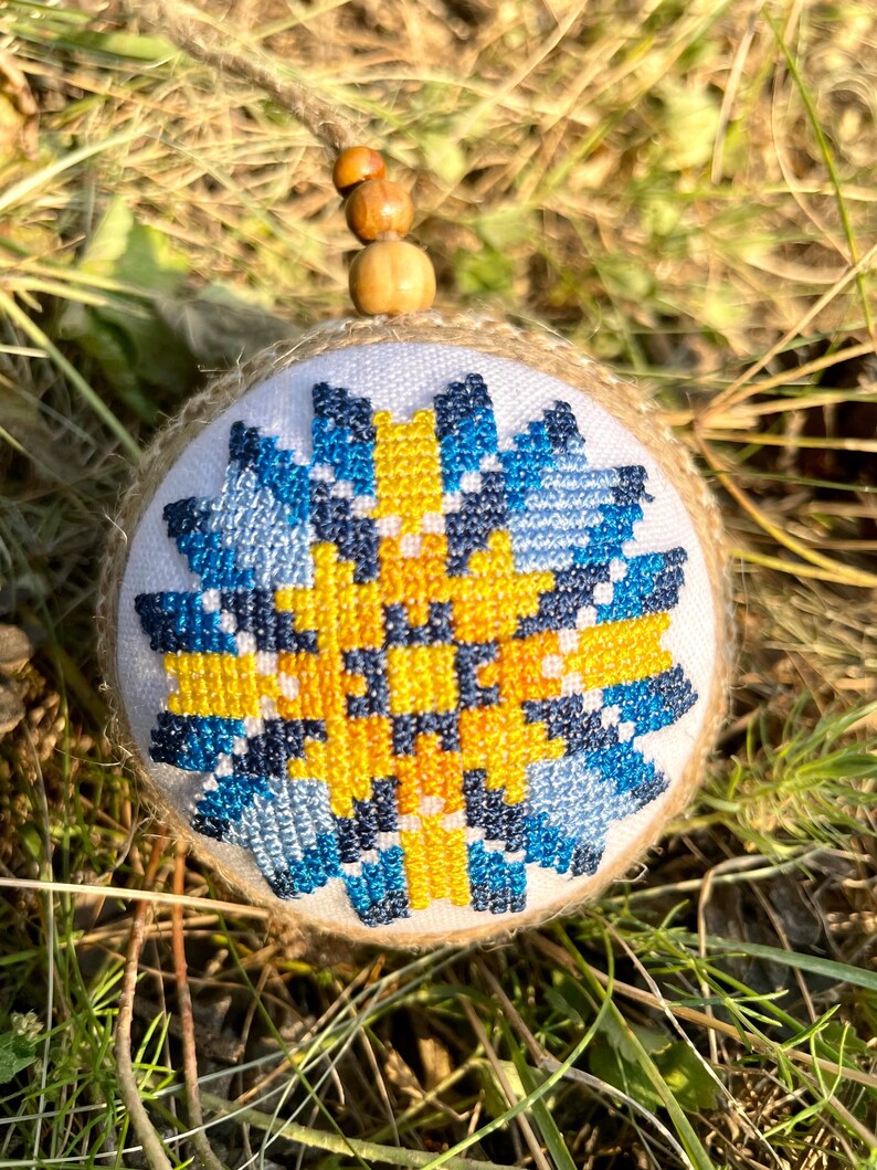 May include: A handmade Christmas ornament with a blue, yellow, and white geometric pattern. The ornament is made of burlap and is decorated with a blue and yellow cross-stitch design.