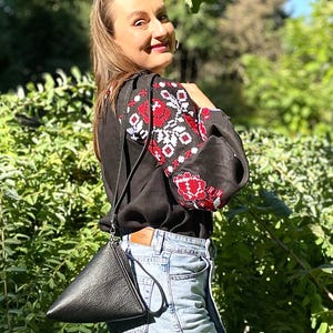 May include: A woman wearing a black linen shirt with red and white embroidery, blue denim shorts, and a black leather triangular bag.