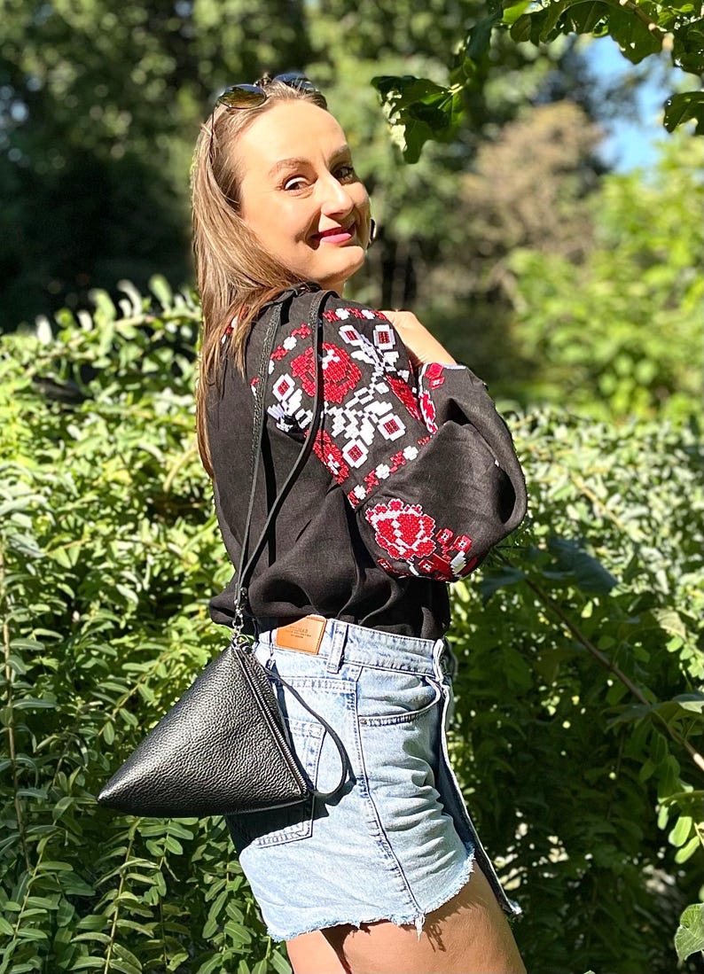May include: A woman wearing a black linen shirt with red and white embroidery, blue denim shorts, and a black leather triangular bag.