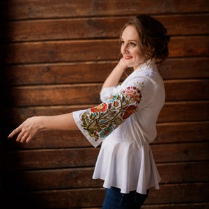 Op de afbeelding: Een vrouw draagt een witte blouse met geborduurde bloemenmotieven op de mouw. De blouse heeft een gerimpelde zoom en ze draagt een blauwe jeans.