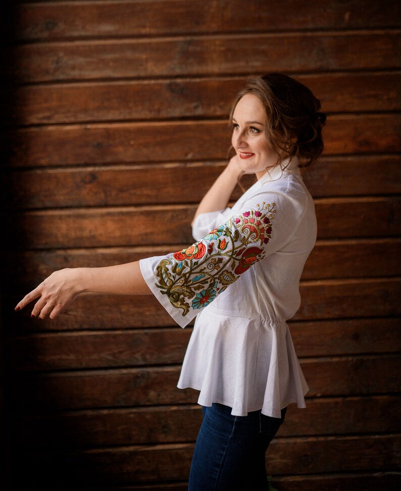 Op de afbeelding: Een vrouw draagt een witte blouse met geborduurde bloemenmotieven op de mouw. De blouse heeft een gerimpelde zoom en ze draagt een blauwe jeans.