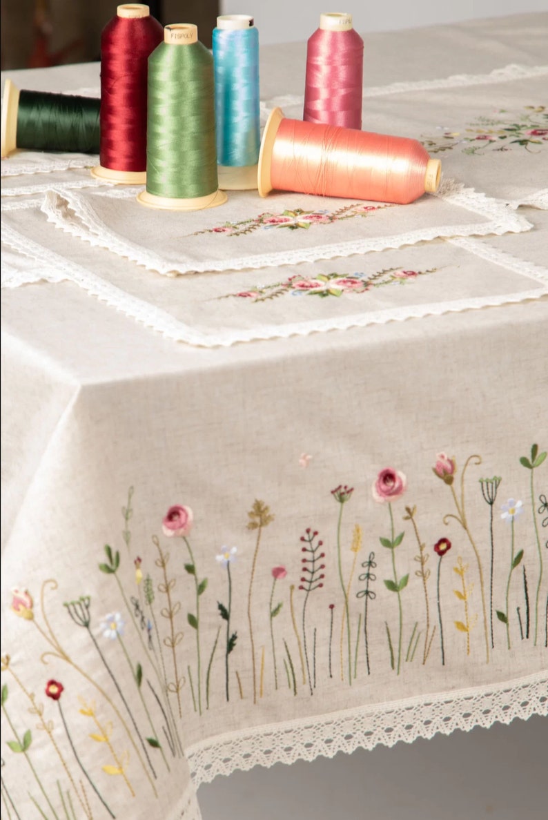 May include: A beige linen tablecloth with embroidered floral designs and lace trim. Colorful spools of thread in various colors, including red, green, and pink, are placed on the tablecloth. The tablecloth features a border of embroidered wildflowers.