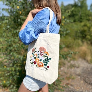 May include: A white canvas tote bag with a floral embroidery design. The embroidery features a variety of flowers in shades of pink, orange, yellow, white, and green.