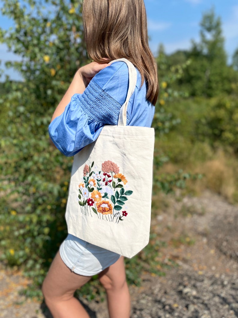 May include: A white canvas tote bag with a floral embroidery design. The embroidery features a variety of flowers in shades of pink, orange, yellow, white, and green.