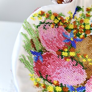 May include: Close-up of a white beaded handbag with a woven handle. The bag features a floral design in shades of pink, green, yellow, and blue. The intricate beadwork creates a textured surface.