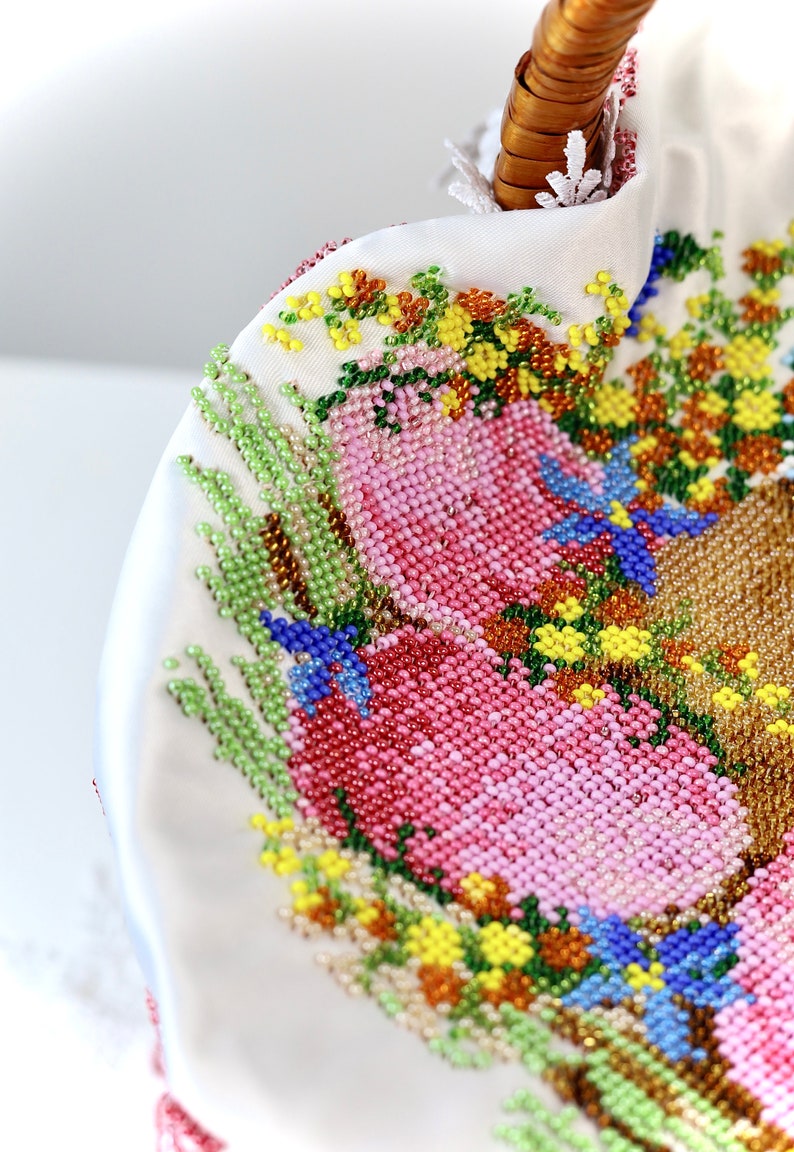 May include: Close-up of a white beaded handbag with a woven handle. The bag features a floral design in shades of pink, green, yellow, and blue. The intricate beadwork creates a textured surface.