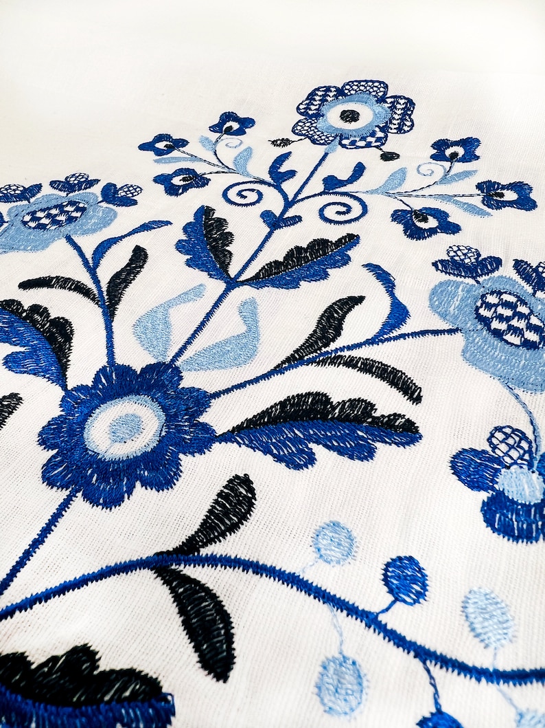 May include: Blue and black embroidered floral design on a white fabric background. The design features stylized flowers and leaves.