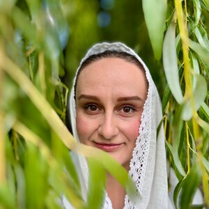 May include: A woman wearing a white head covering with a delicate lace trim, framed by green willow tree leaves. The woman has brown eyes and a slight smile, with a natural makeup look. The background is blurred, creating a soft focus.