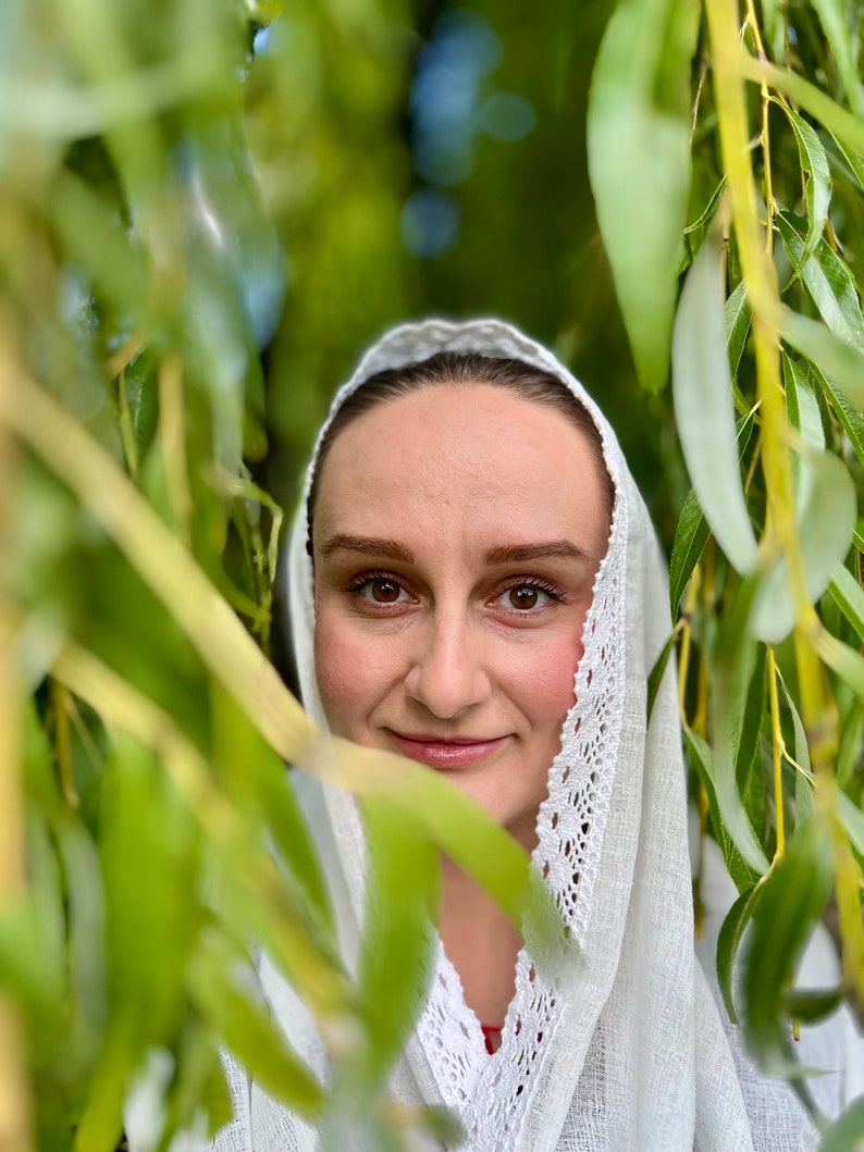 May include: A woman wearing a white head covering with a delicate lace trim, framed by green willow tree leaves. The woman has brown eyes and a slight smile, with a natural makeup look. The background is blurred, creating a soft focus.