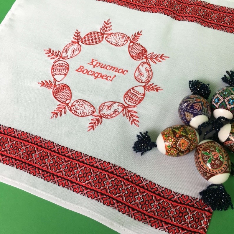 May include: White embroidered table runner with a red embroidered wreath of eggs and the text "Христос Воскрес!" in red. The table runner has a red and black border. There are several decorated Easter eggs on the table runner.