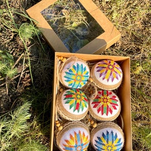 May include: Six handmade Christmas ornaments in a brown box. The ornaments are round and covered in burlap. Each ornament features a colorful embroidered star design.