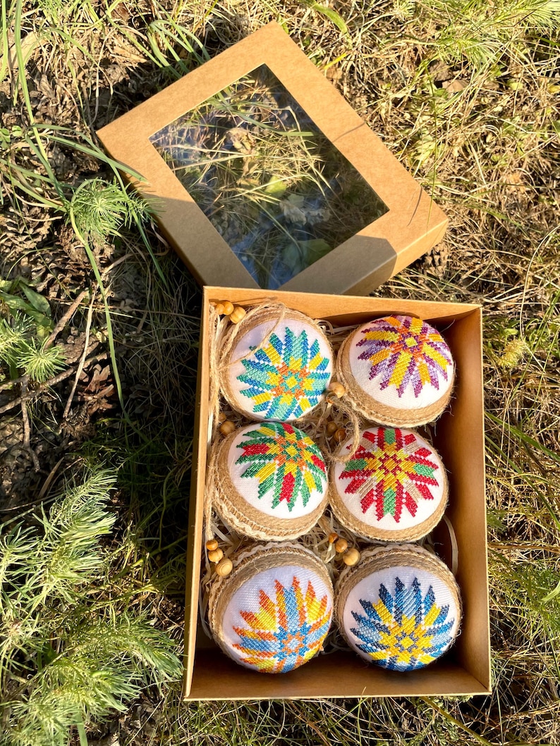 May include: Six handmade Christmas ornaments in a brown box. The ornaments are round and covered in burlap. Each ornament features a colorful embroidered star design.