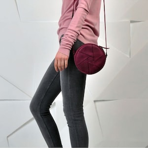May include: A burgundy-colored, round crossbody bag with a geometric design on the front. The bag has a long, thin strap and a zipper closure. The bag is shown against a white background.