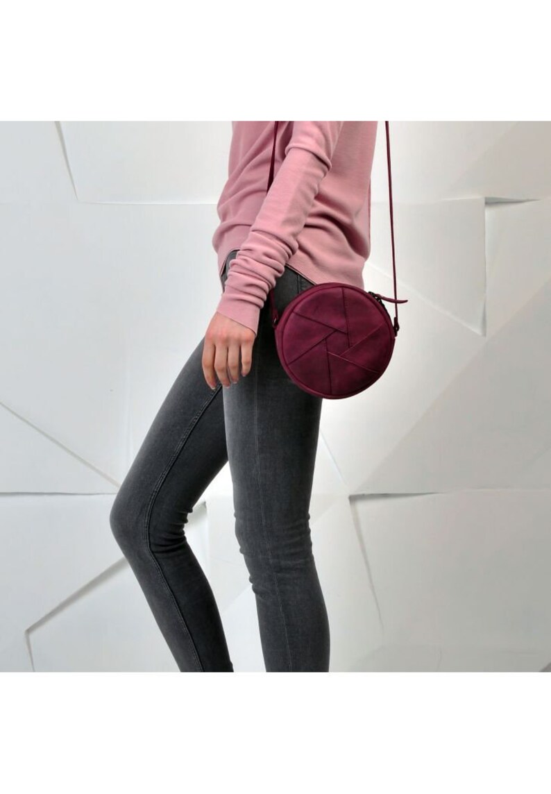 May include: A burgundy-colored, round crossbody bag with a geometric design on the front. The bag has a long, thin strap and a zipper closure. The bag is shown against a white background.
