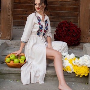 Pu&ograve; includere: Una donna con un vestito bianco di lino con ricami colorati siede su una serie di gradini di pietra. Sta tenendo un cesto di mele verdi. Ci sono fiori rossi e gialli su entrambi i lati di lei.