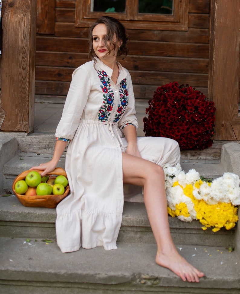 Pu&ograve; includere: Una donna con un vestito bianco di lino con ricami colorati siede su una serie di gradini di pietra. Sta tenendo un cesto di mele verdi. Ci sono fiori rossi e gialli su entrambi i lati di lei.